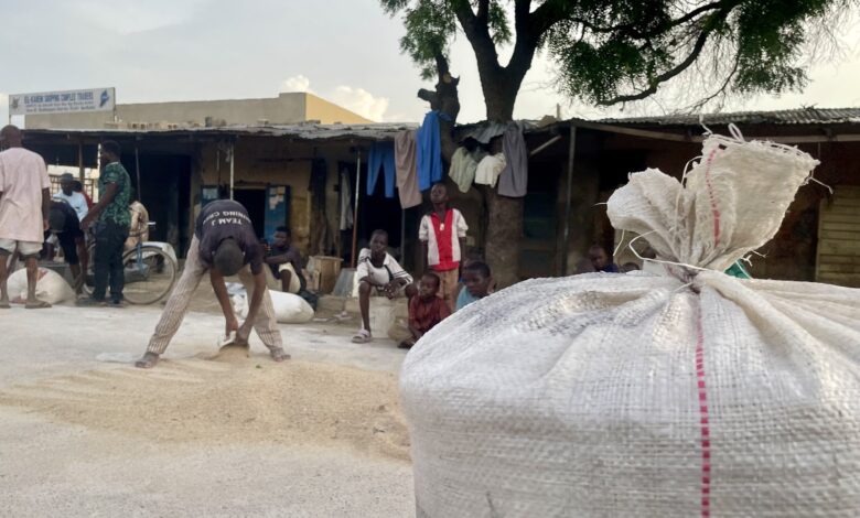 People in a market with large bags in the foreground and a tree providing shade.