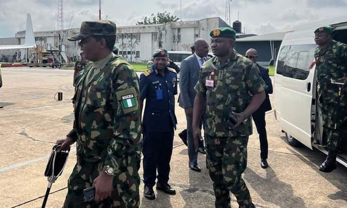 Military personnel walking on a tarmac with a white van and building in the background.