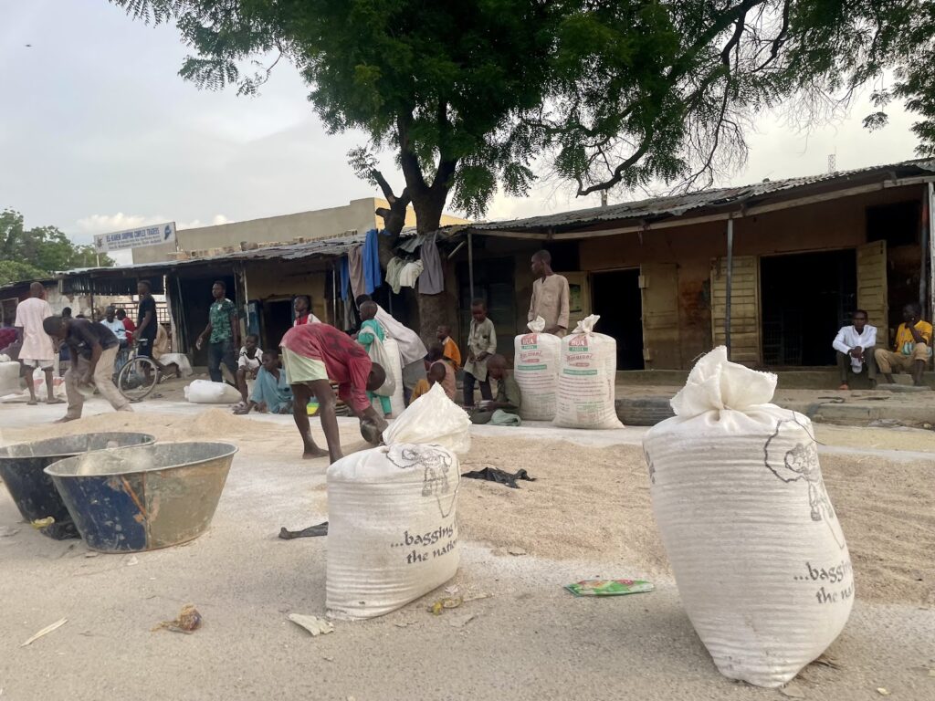 Street scene with people sitting and working near large bags and a basin under a tree, with a storefront in the background.