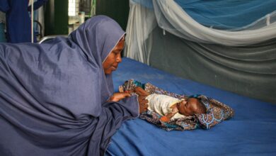 Woman in a blue hijab looking at a baby lying on a hospital bed, under a mosquito net in a medical facility.