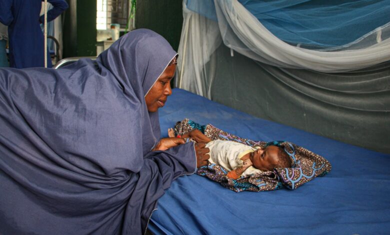 Woman in a blue hijab looking at a baby lying on a hospital bed, under a mosquito net in a medical facility.