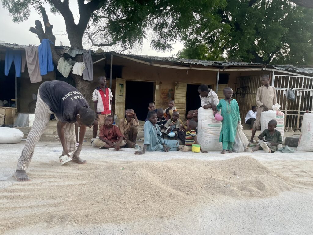 A group of people, including children, gathering around piles of grain on the ground in front of a building with hanging laundry.