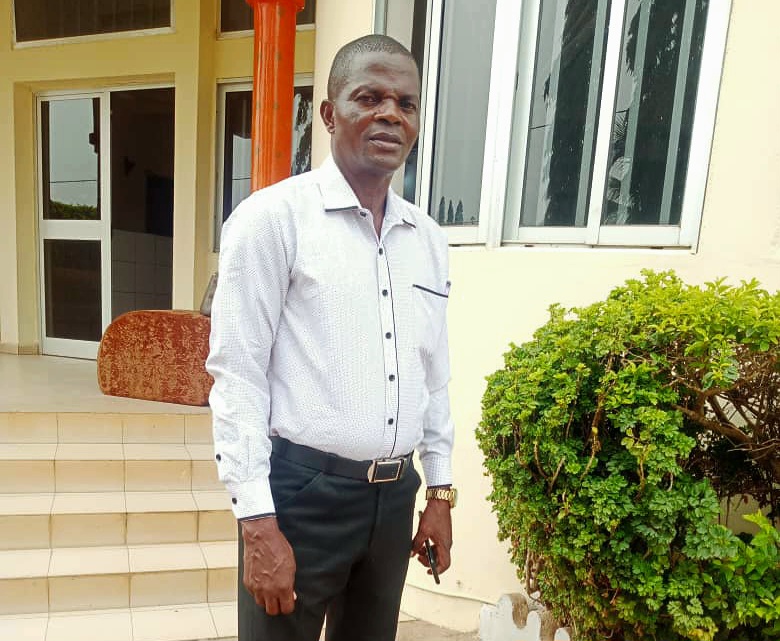 Man in a white shirt and black pants standing in front of a building with a bush.