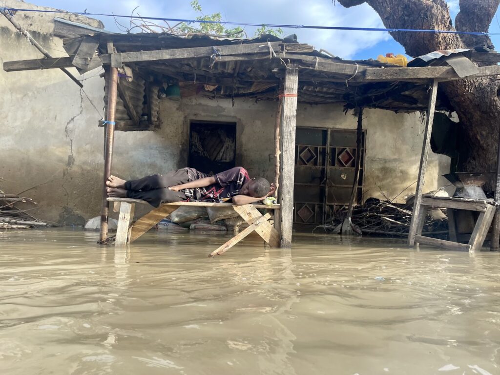 Person lying on makeshift bed above floodwater near a damaged house.