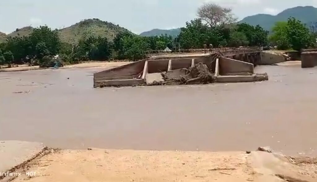 Floodwaters surrounding and damaging a bridge with trees and hills in the background.