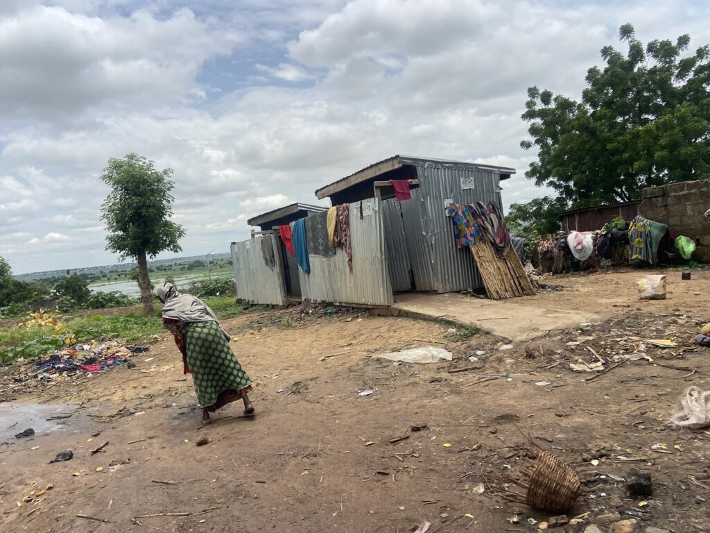 A woman walks by makeshift houses with clothes hanging outside under a cloudy sky.