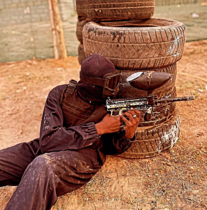 Person in tactical gear aiming a paintball gun from behind stacked tires.