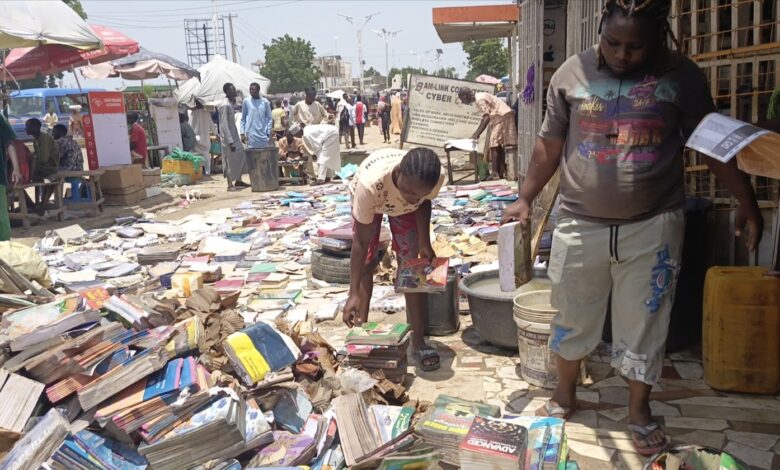 Street book market with people browsing piles of books on the ground under the sun.