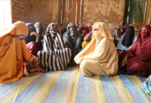 Group of women in colorful attire sitting on a striped mat indoors, with sunlight streaming in.