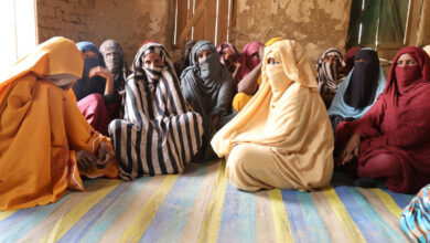 Group of women in colorful attire sitting on a striped mat indoors, with sunlight streaming in.