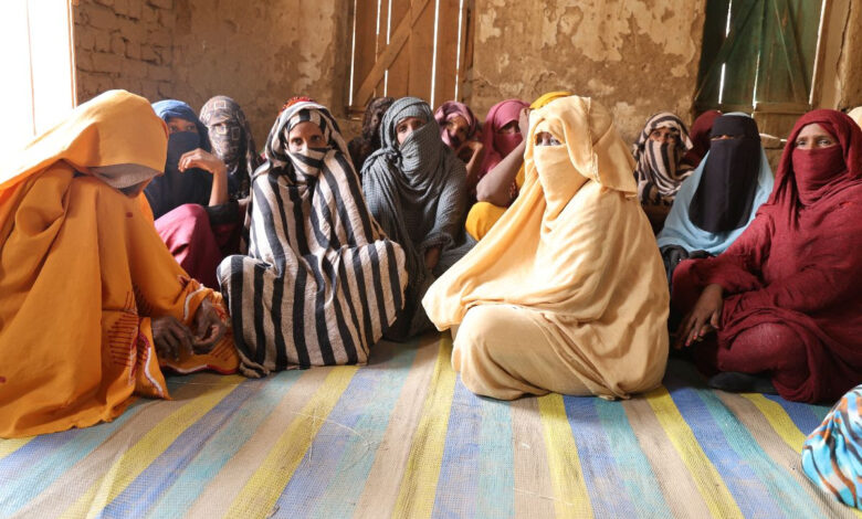 Group of women in colorful attire sitting on a striped mat indoors, with sunlight streaming in.