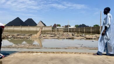 A man in traditional clothing stands beside flooded ruins with houses in the background.