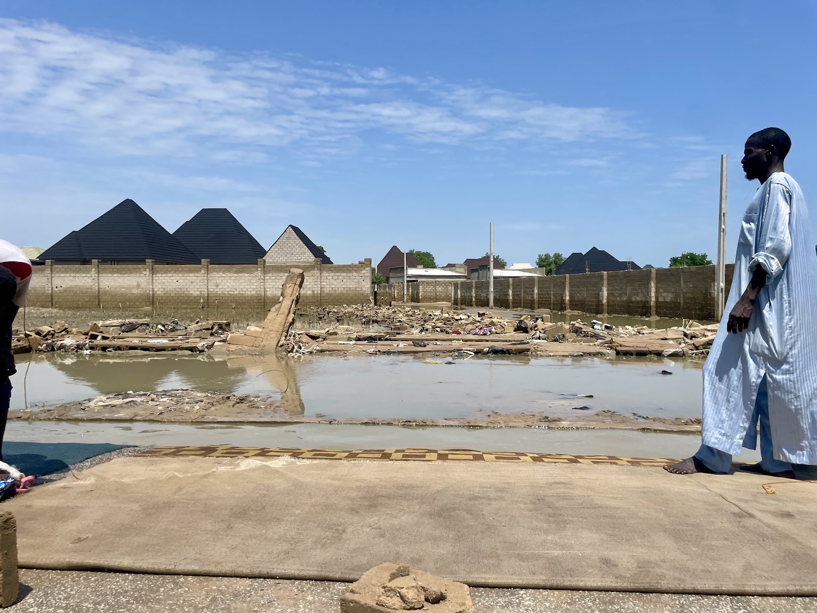 A man in traditional clothing stands beside flooded ruins with houses in the background.