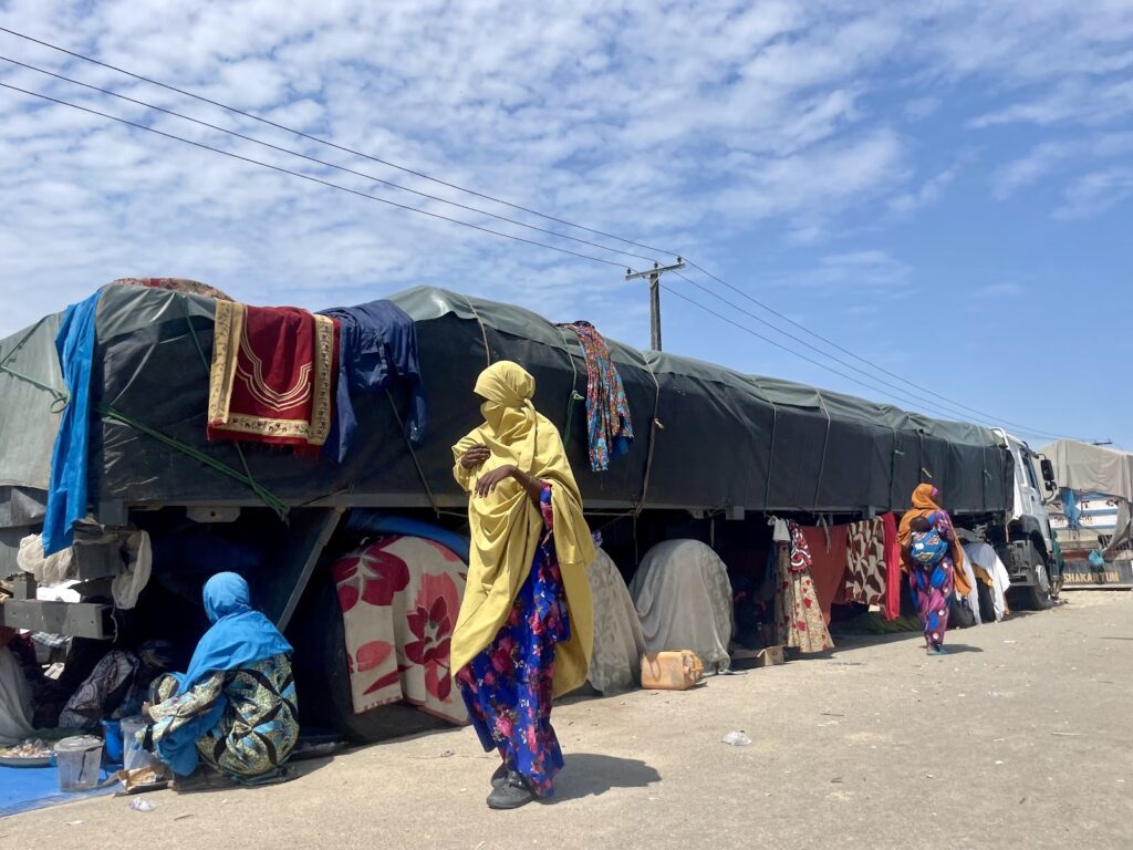 Women near a covered truck with colorful garments in a market setting under a clear sky.