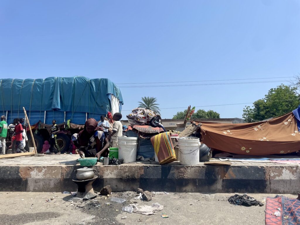 Busy outdoor market scene with people, covered goods, and a person cooking on a small stove.