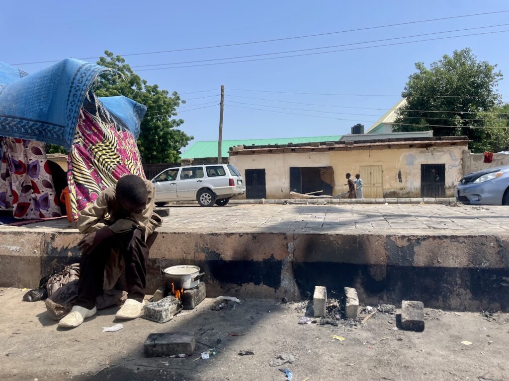 Person sitting by a small fire on the street with colorful fabric and buildings in the background.