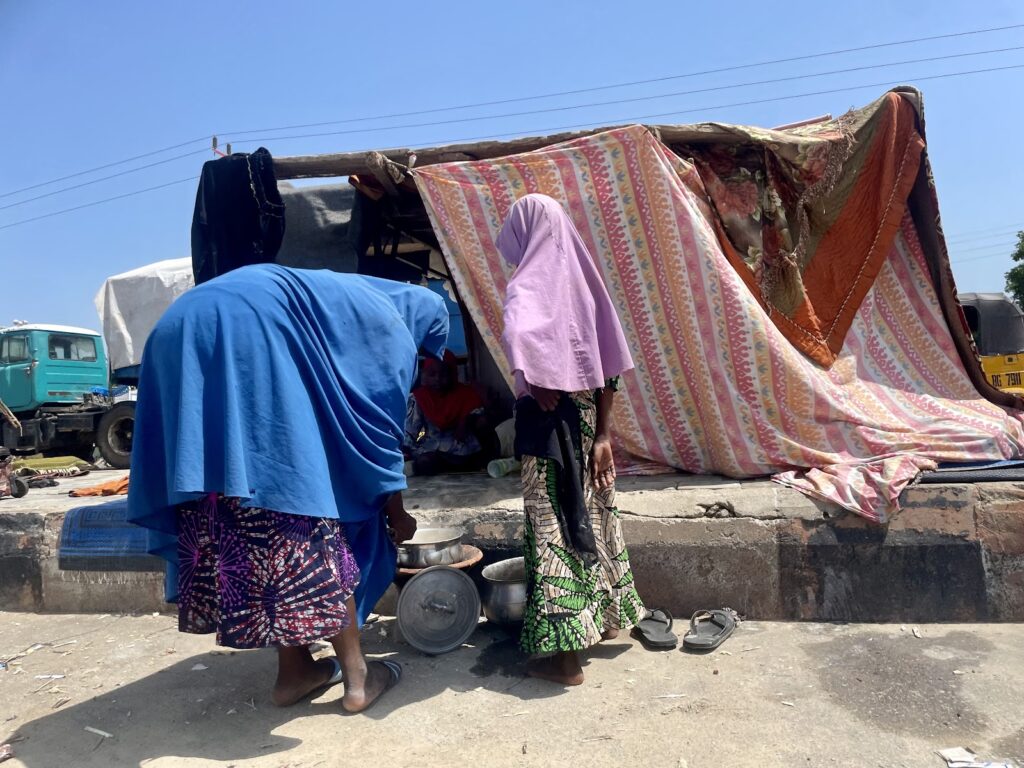 Two people outside a makeshift tent on a sunny day, with a glimpse of urban life in the background.