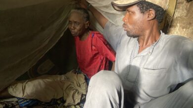 Two people sitting under a makeshift tent, one looking weary and the other gazing into the distance.