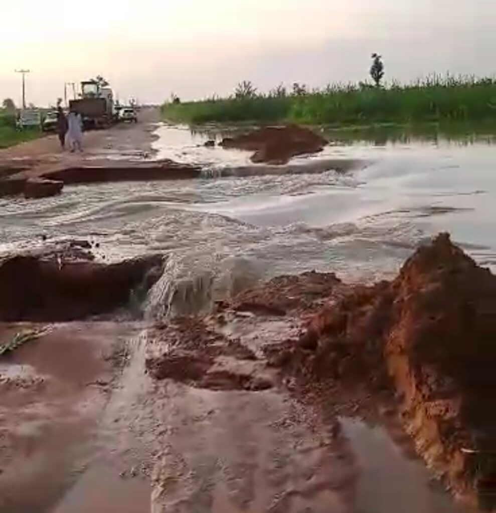 A damaged road with water flowing through, creating a large erosion gap as a person observes nearby.