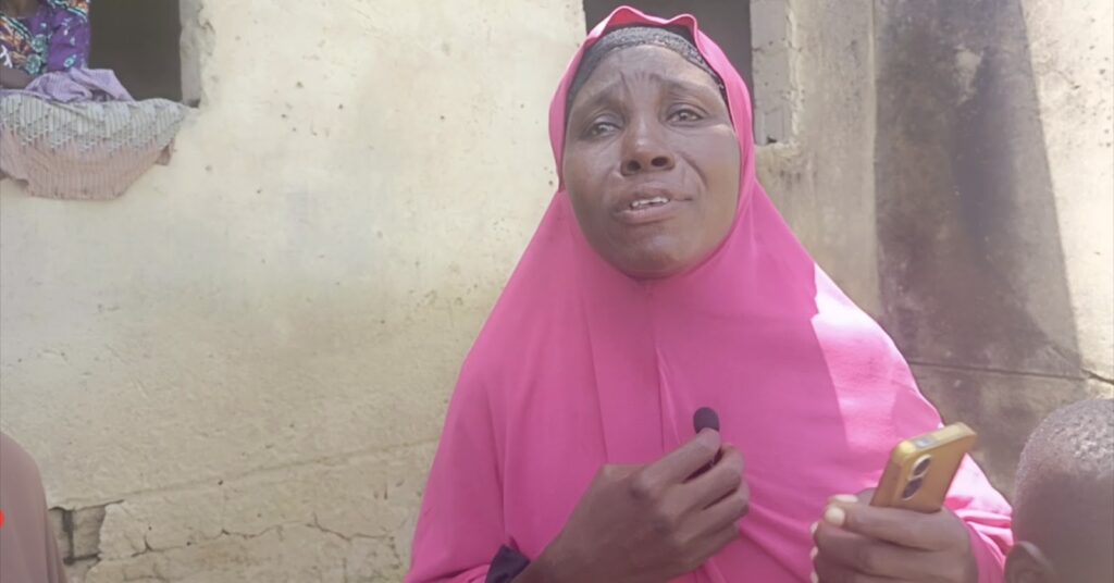 Woman in pink hijab speaking, holding a microphone, with a smartphone in her other hand, standing against a weathered wall.