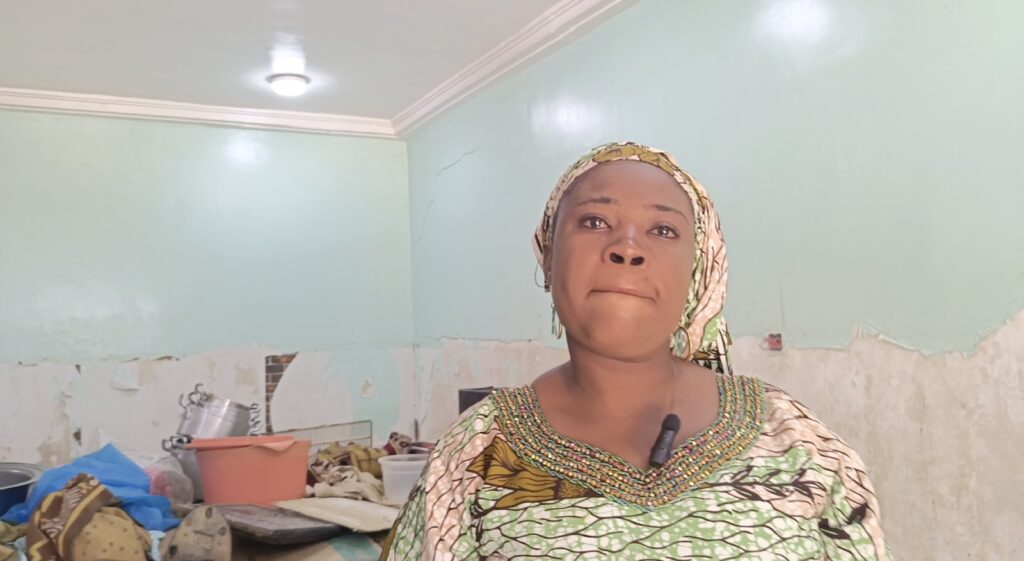 Woman in patterned clothing standing in a room with peeling walls and household items in the background.