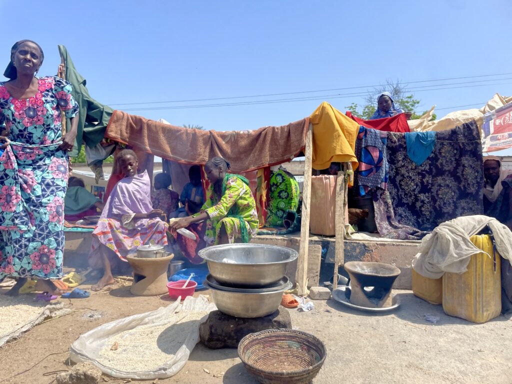 People at an outdoor market with colorful clothing and textiles, cooking pots and baskets on the ground.