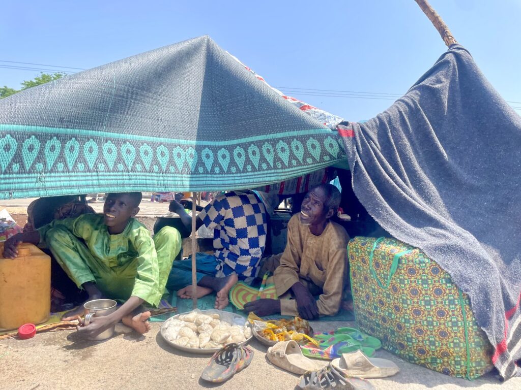 People sitting under a makeshift tent with a cloth spread of goods, surrounded by personal items.