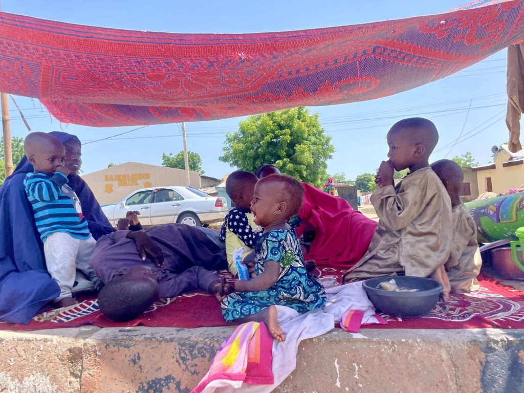 Family sitting on a mat under a shade cloth, with children playing and a man holding a baby.