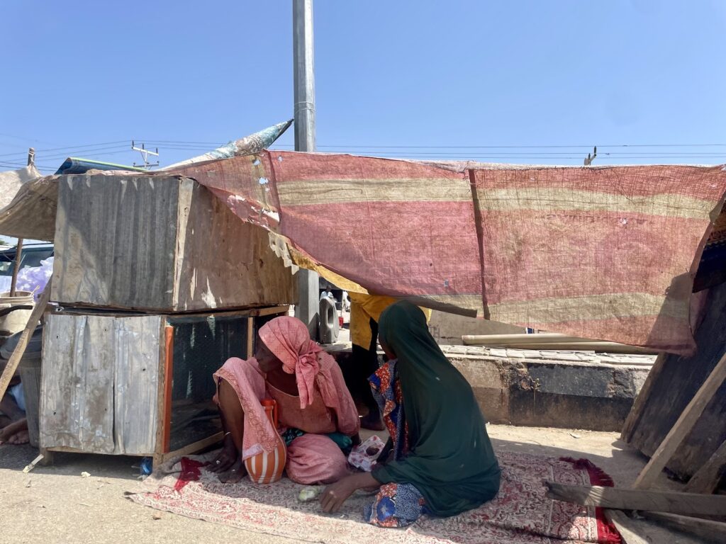 Two individuals sitting under a makeshift shelter with corrugated metal and tarp on a sunny day.