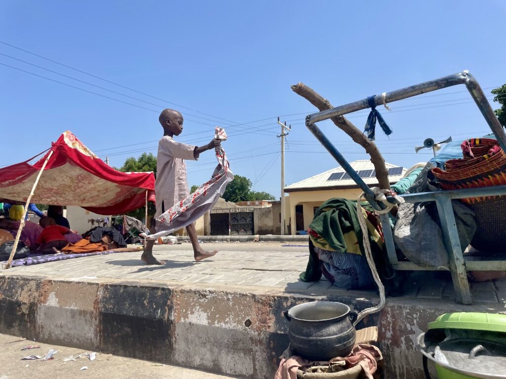 A young child walks barefoot carrying a piece of cloth, with street vendors and their wares under makeshift tents in the background.