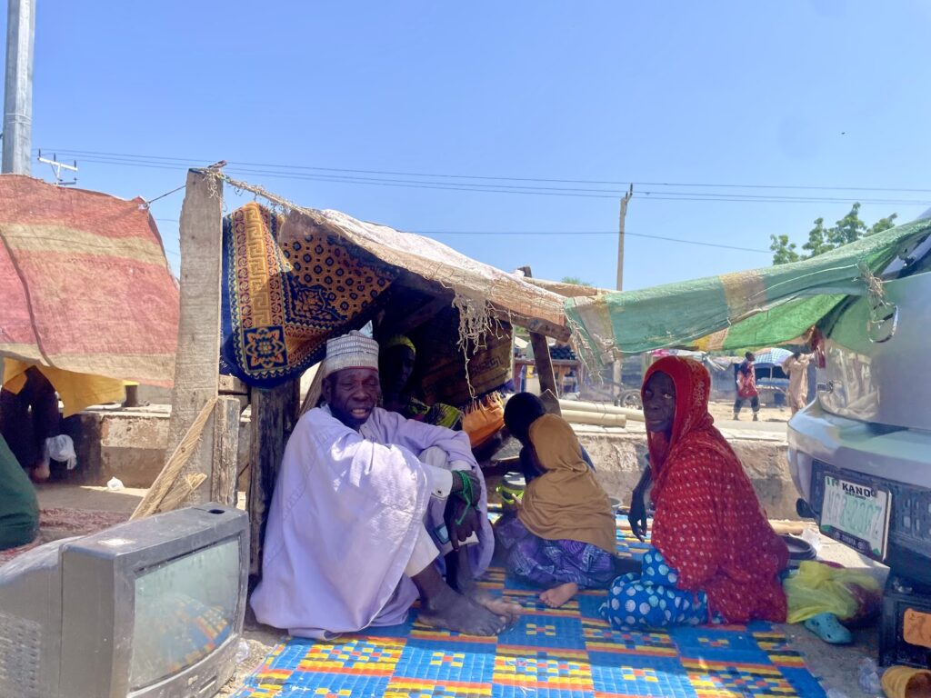 People sitting under makeshift tents with colorful fabrics in a sunny market setting.