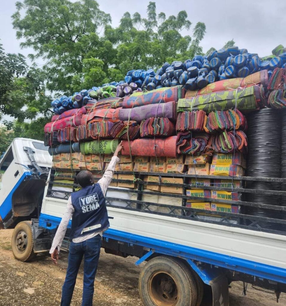 Person gesturing to a truck heavily loaded with colorful mats and black rolls, surrounded by greenery.