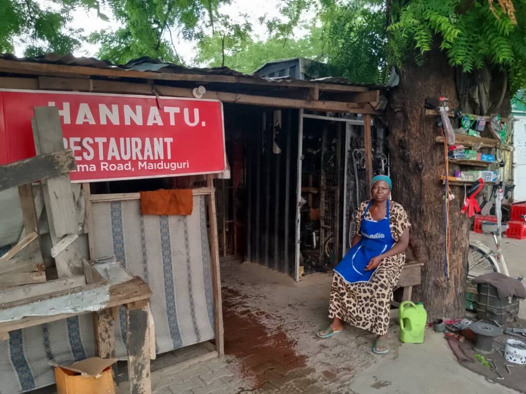 Woman in blue apron seated next to a roadside restaurant under a red signboard, with a large tree and various items around.