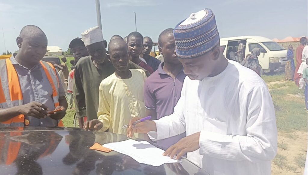 Man in white traditional attire signing a document on a car hood with onlookers in the background.