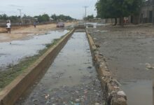 Flooded street with trash-strewn water and a red car driving through, with people walking on the sides.