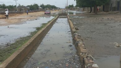 Flooded street with trash-strewn water and a red car driving through, with people walking on the sides.