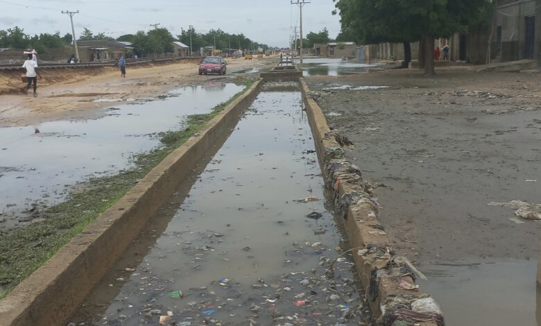 Flooded street with trash-strewn water and a red car driving through, with people walking on the sides.