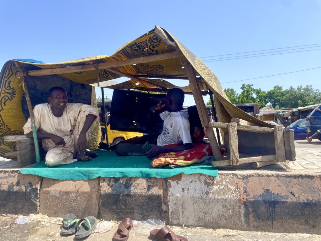 Two people relaxing under a makeshift shelter by a roadside on a sunny day.