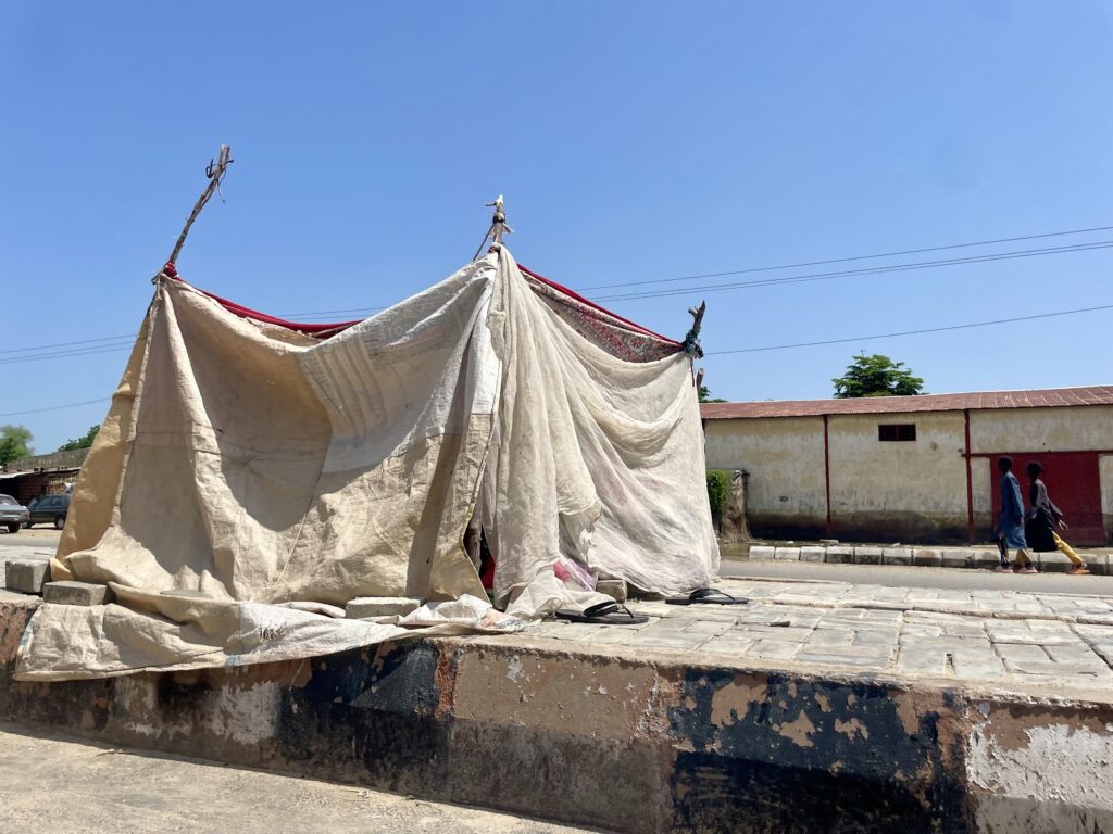 A makeshift tent constructed from tattered fabric on a sunny day with two people walking in the background.