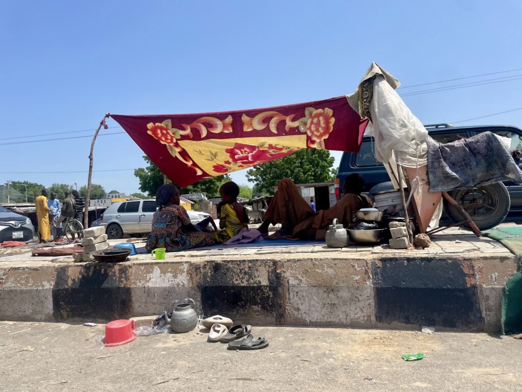 People sitting under a makeshift tent on a sunny day, with cooking utensils and bicycles nearby.