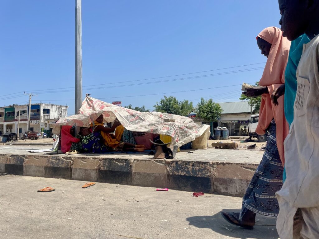 People walking by a makeshift street shelter under a fabric canopy, in a sunny, bustling environment.