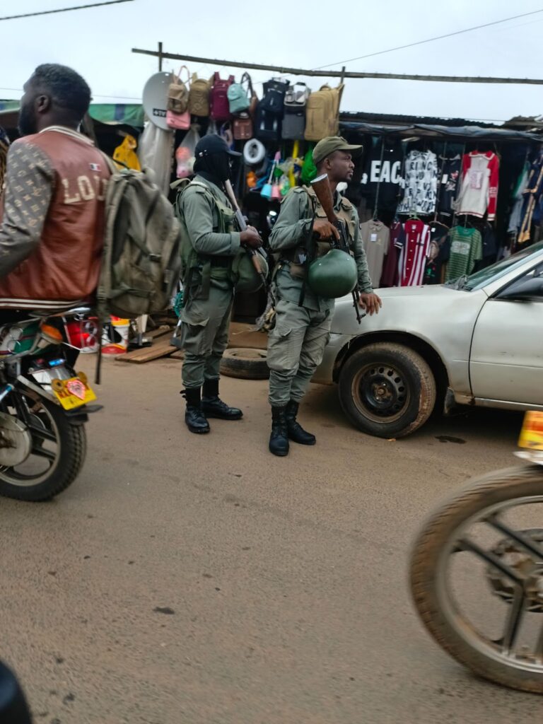 Two armed personnel in uniform patrolling a busy street market with shops and vehicles.