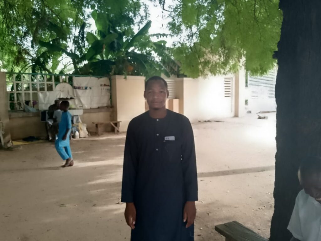 Man in traditional attire standing in a courtyard with trees and a building in the background.