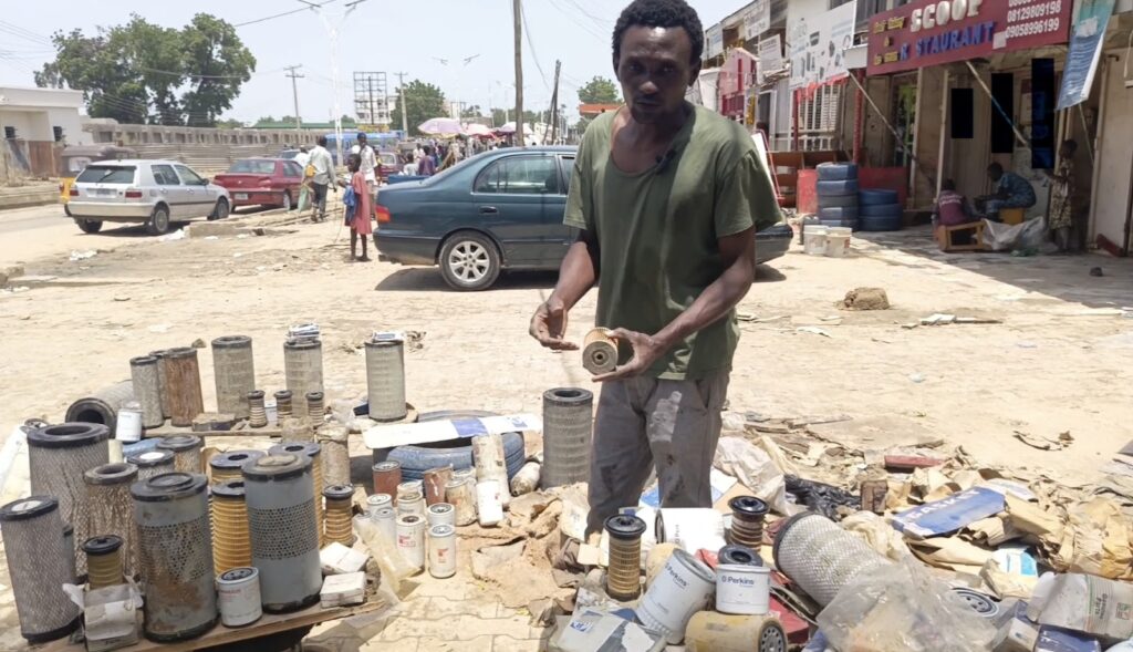 Man selling various used car filters on a street with onlookers and vehicles in the background.