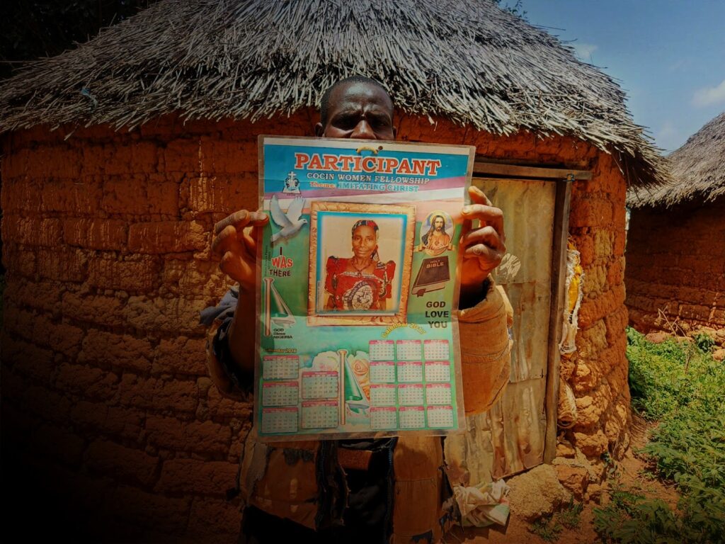 A person holding a colorful religious poster in front of a thatched-roof mud house.