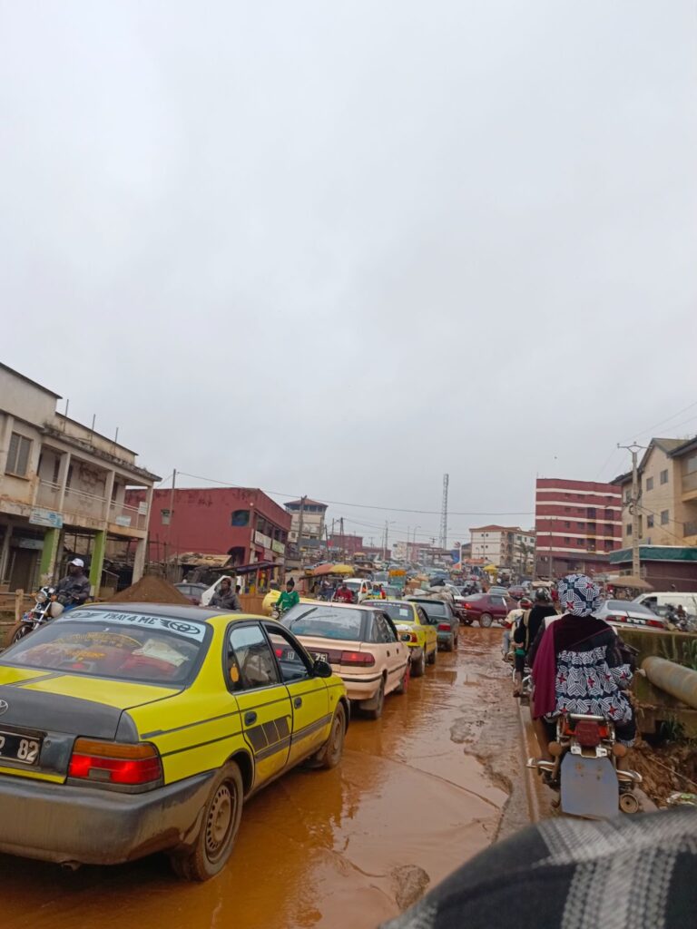 Busy street with cars and motorcycles navigating through a muddy road amidst market activity under an overcast sky.