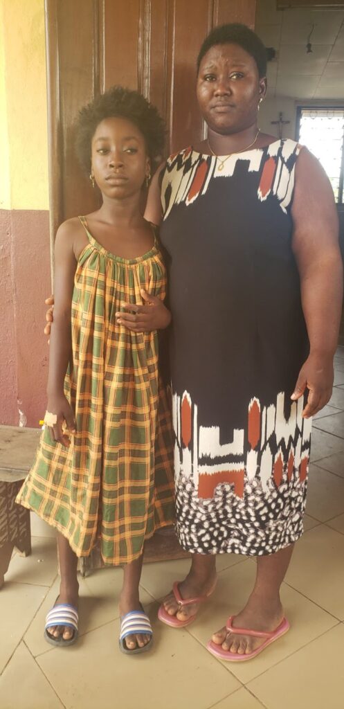 Two people standing side by side, one young, one older, in patterned clothing and sandals.