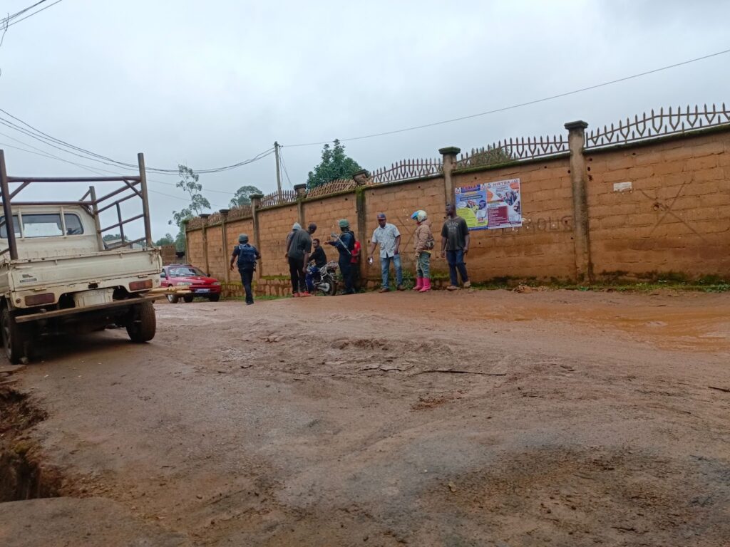 People standing by a muddy road with vehicles and a wall with a poster.