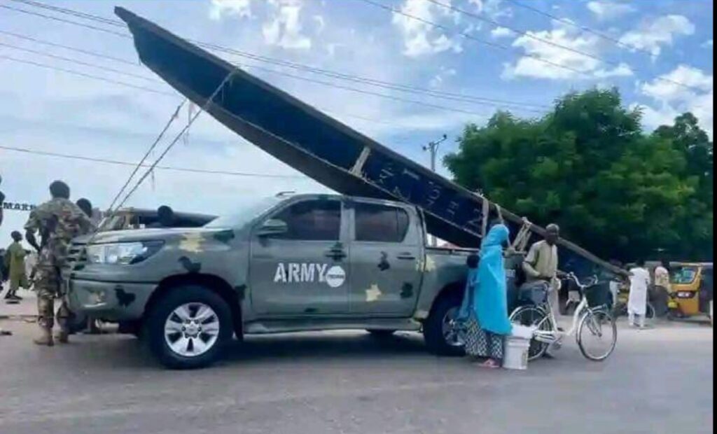 Army vehicle with a canoe strapped on top at a busy intersection with soldiers and civilians nearby.