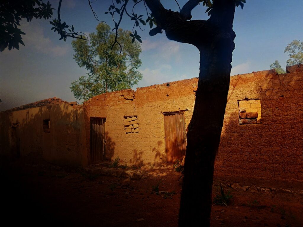 Old mud-brick house with a wooden door and windows, tree in foreground under a hazy sky.